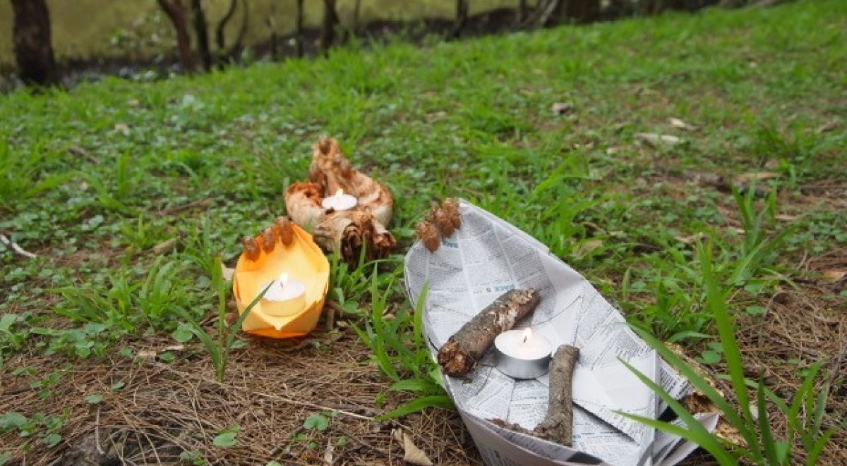 Tea light candles inside paper boats laying on grass near the banks of the river