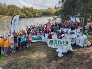 Large group of community members standing beside the river with hands in the air, pictured behind collected bags of rubbish and range of community group banners including River Canoe Club, Mudcrabs and Cooks River Valley Association