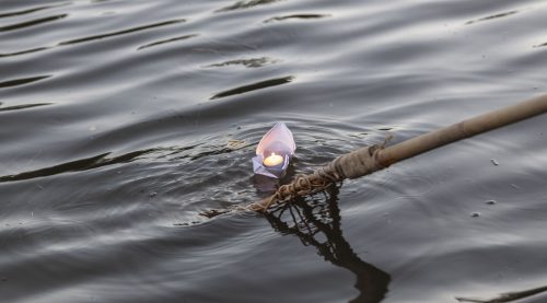 An image of a wooden paddle releasing a paper boat into the water. Inside the paper boat is a candle.